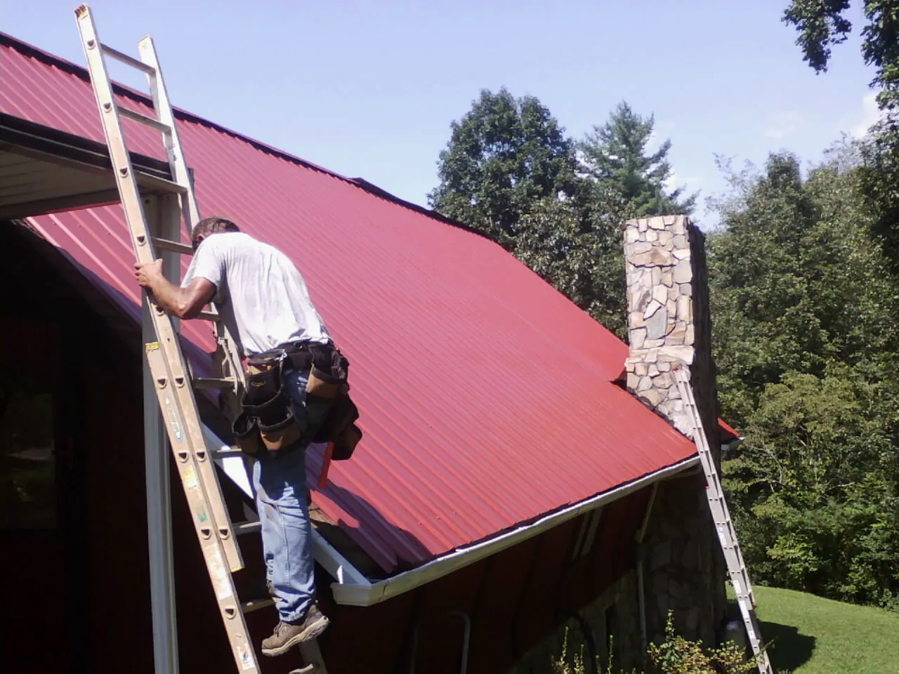Roofer on a ladder beside a red metal roof and stone chimney — Soffit & Fascia Repair by Roofing Brothers in Snook